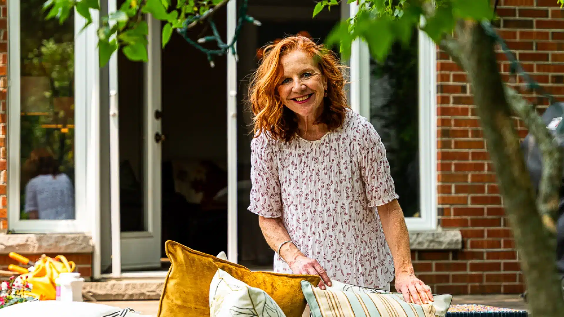 smiling woman with curly red hair stands by a sofa, arranging cushions in a backyard setting.
